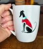 White mug with a greyhound  illustration wearing a red Christmas sweater held by a hand on a wooden surface.