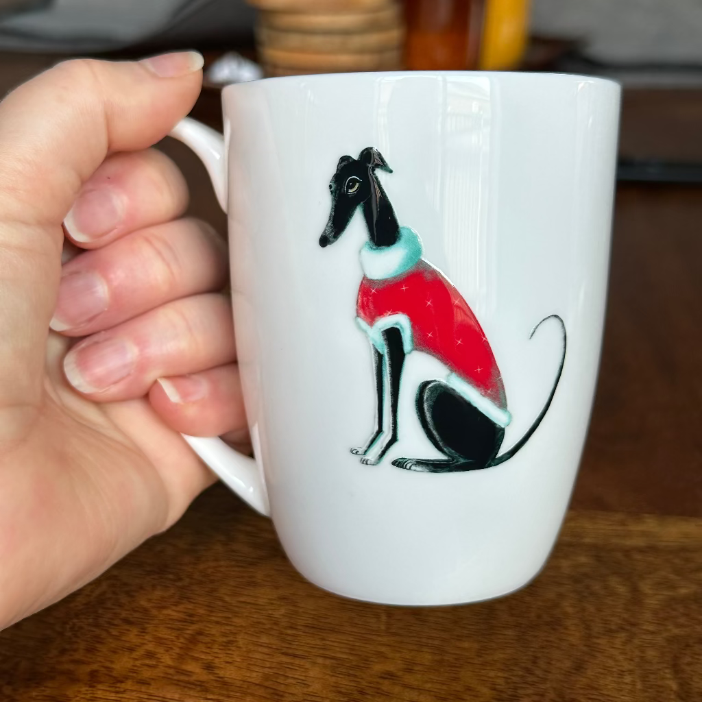 White mug with a greyhound  illustration wearing a red Christmas sweater held by a hand on a wooden surface.