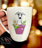 White mug with a whippet  illustration in a pot of flowers, held by a hand against a blurred background.
