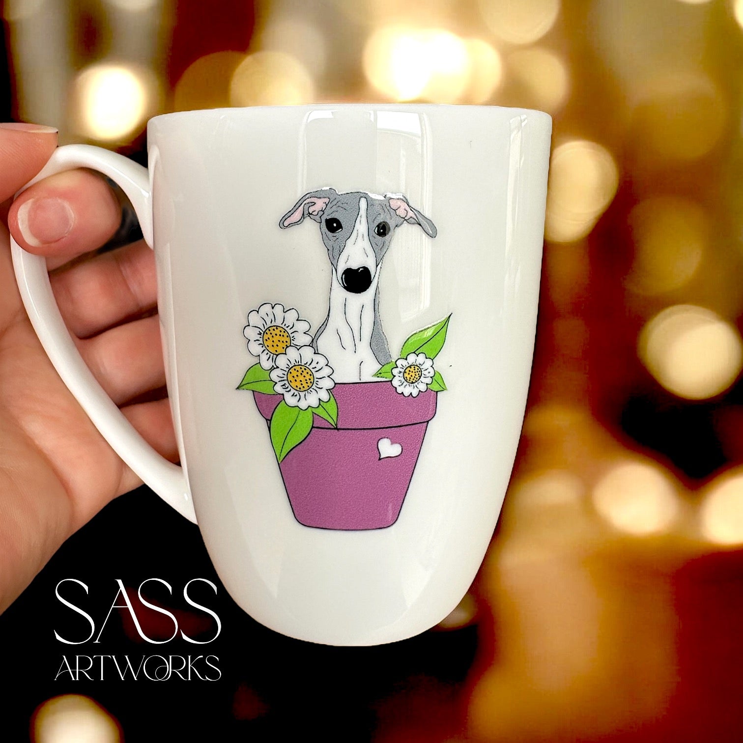 White mug with a whippet  illustration in a pot of flowers, held by a hand against a blurred background.