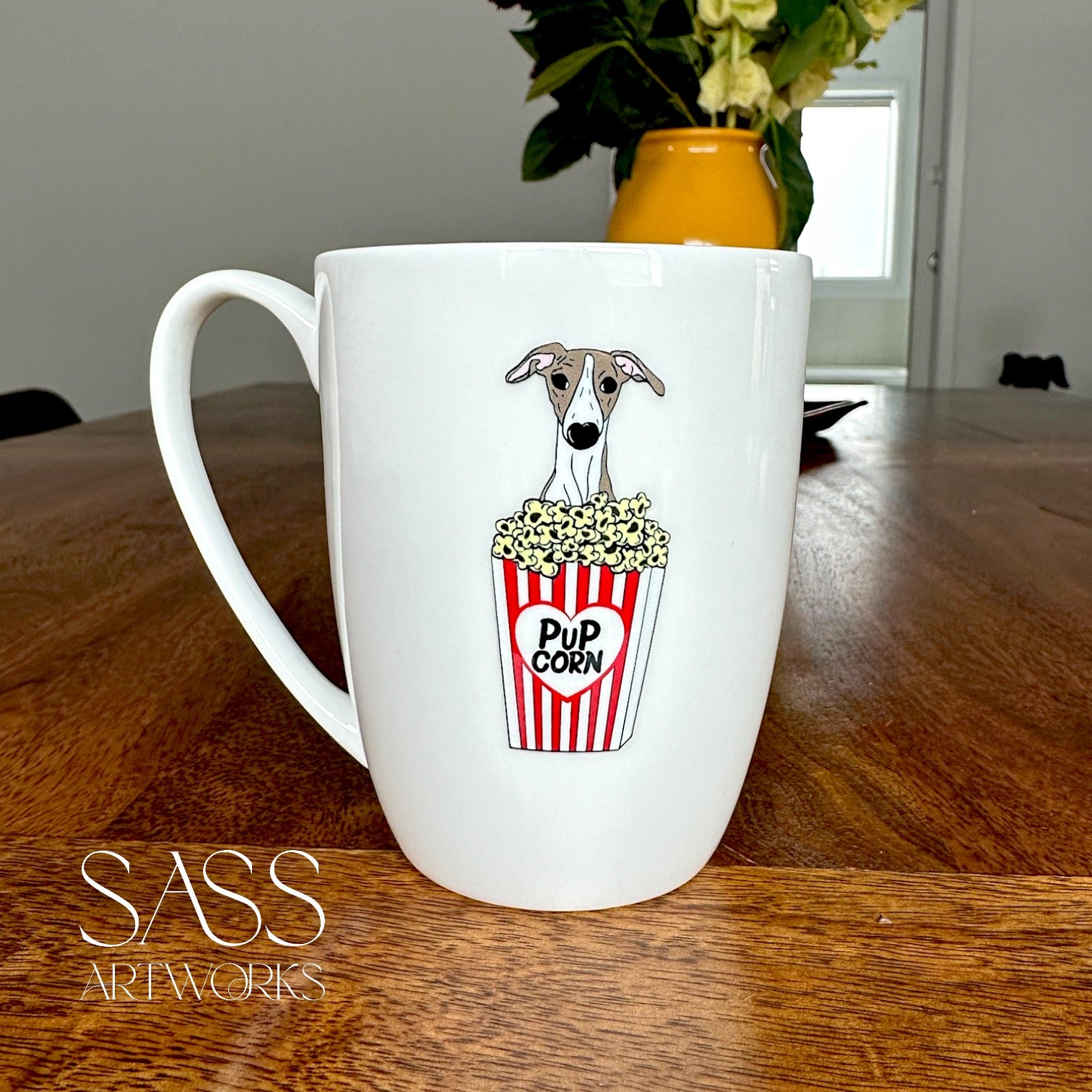 White mug with a whippet and popcorn design on a wooden table, featuring 'SASS Artworks' branding.
