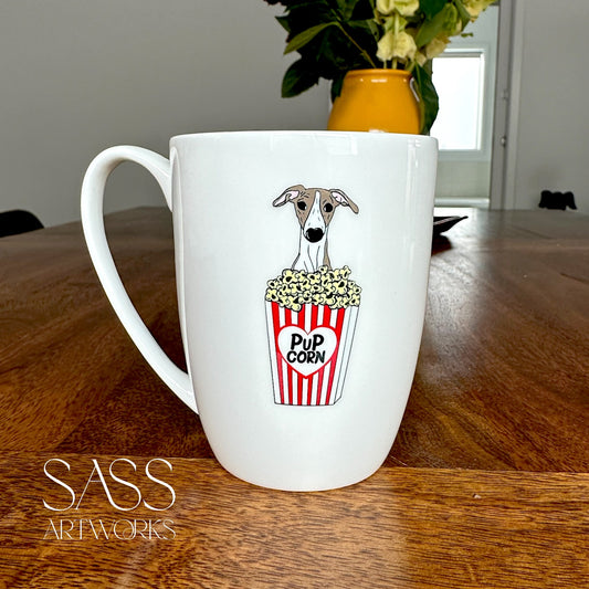 White mug with a whippet and popcorn design on a wooden table, featuring 'SASS Artworks' branding.
