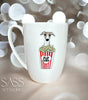 White mug with a whippet illustration holding popcorn, set against a blurred bokeh background. Brand 'SASS Artworks' visible.