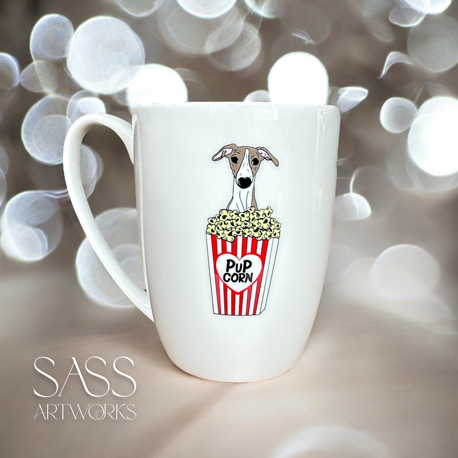 White mug with a whippet illustration holding popcorn, set against a blurred bokeh background. Brand 'SASS Artworks' visible.