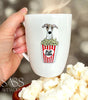 White mug with a whippet illustration holding popcorn, held by a hand with popcorn in the foreground.