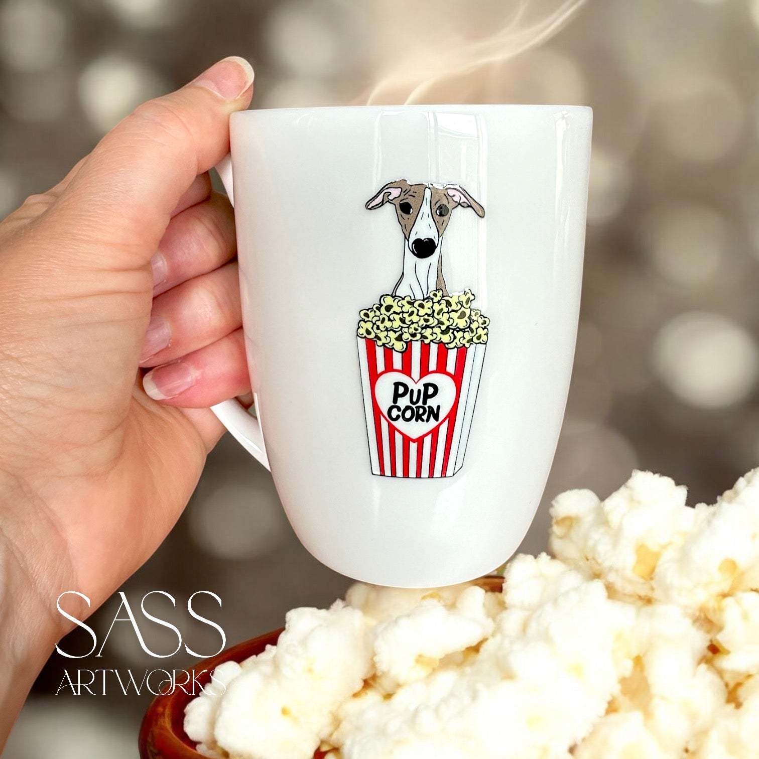 White mug with a whippet illustration holding popcorn, held by a hand with popcorn in the foreground.