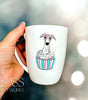 White mug with a whippet  in a cupcake design held by a hand, blurred lights in the background