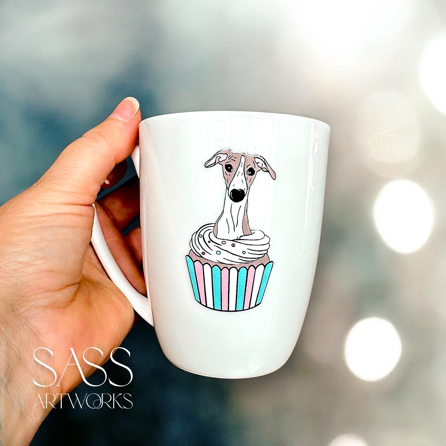 White mug with a whippet  in a cupcake design held by a hand, blurred lights in the background