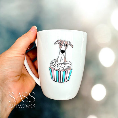 White mug with a whippet  in a cupcake design held by a hand, blurred lights in the background