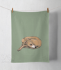 Green tea towel with a whippet illustration hanging on a line against a white wall.